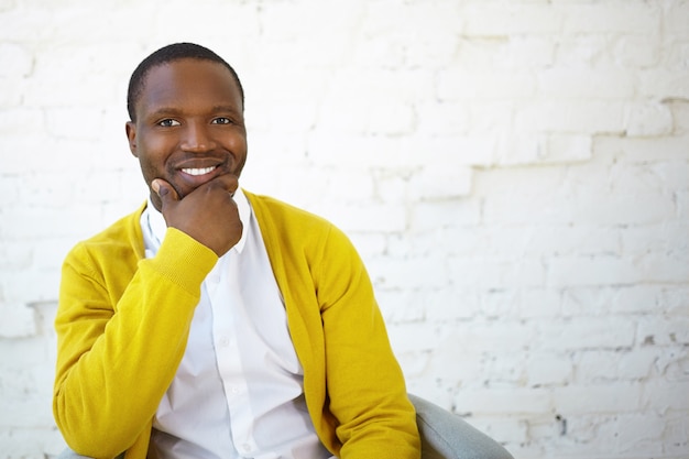 Foto grátis foto de um homem negro e atraente, alegre, com um casaco de lã amarelo, segurando a mão no queixo e sorrindo amplamente para a câmera, sentindo-se feliz ou inspirado, alegrando-se com boas notícias positivas, posando no estúdio