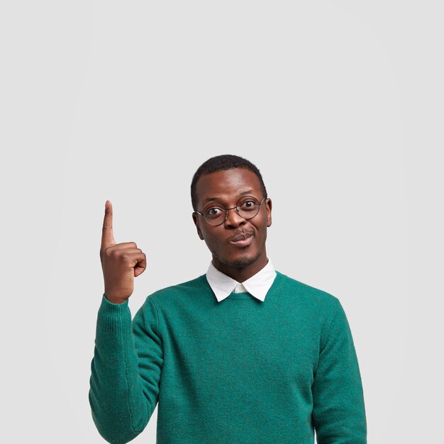 Foto de um homem negro, indeciso, franzindo os lábios, levanta o dedo da frente e fica de pé sobre uma parede branca