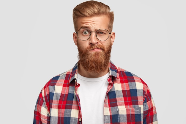 Foto de um homem perplexo e perplexo com espessa barba ruiva e bigode, levanta sobrancelhas, parece duvidoso, usa roupas da moda, isolado sobre uma parede branca. Expressões faciais