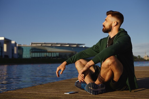 Foto de um jovem caucasiano com barba por fazer e tênis, sentado de pernas cruzadas na calçada de madeira à beira do lago, meditando, ouvindo música calma usando o aplicativo gratuito em seu dispositivo eletrônico