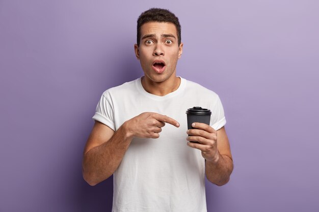 Foto de um jovem estupefato apontando o dedo para o café para viagem, surpreso por ter o copo de papel vazio sem qualquer bebida, usa uma camisa casual branca, abriu a boca, isolado contra a parede roxa.