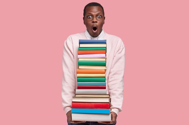 Foto de um jovem negro espantado com um macacão branco casual carregando uma pilha de livros, usando óculos redondos e encostada na parede rosa