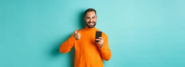 Foto de um jovem satisfeito com um suéter laranja mostrando o polegar para cima depois de ler no telefone móvel