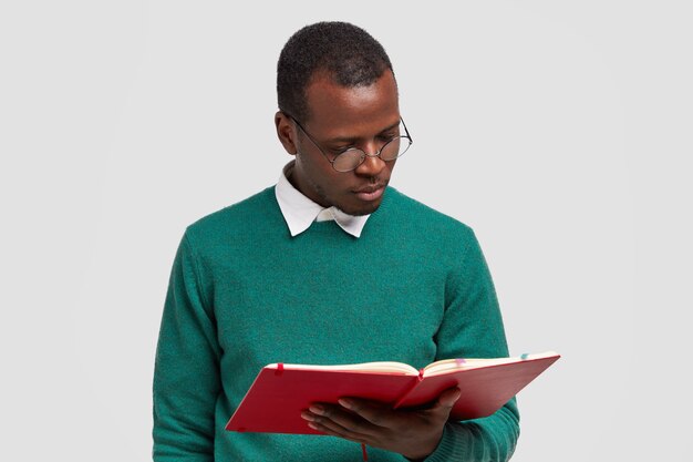 Foto de um jovem sério de pele escura concentrado em um livro didático, usando óculos redondos, suéter verde, estudando na faculdade