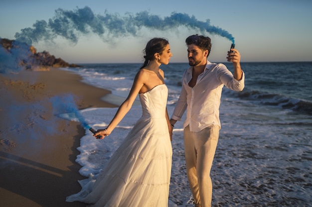 Foto de um lindo casal posando com uma bomba de fumaça azul na praia