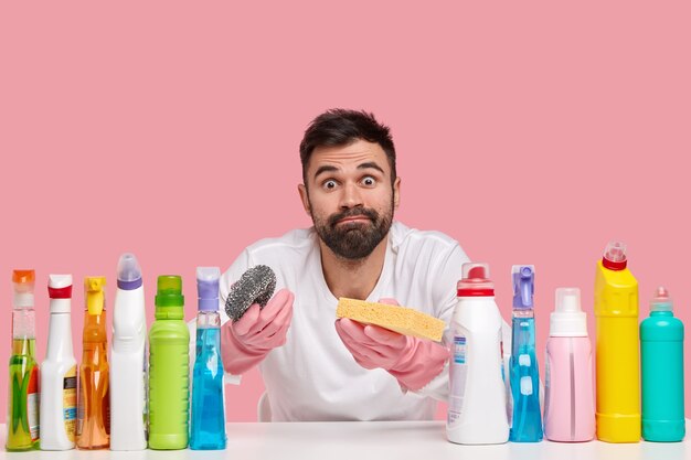 Foto de um marido barbudo atraente e ocupado segurando duas esponjas, erguendo as sobrancelhas surpreso, usando luvas de proteção de borracha