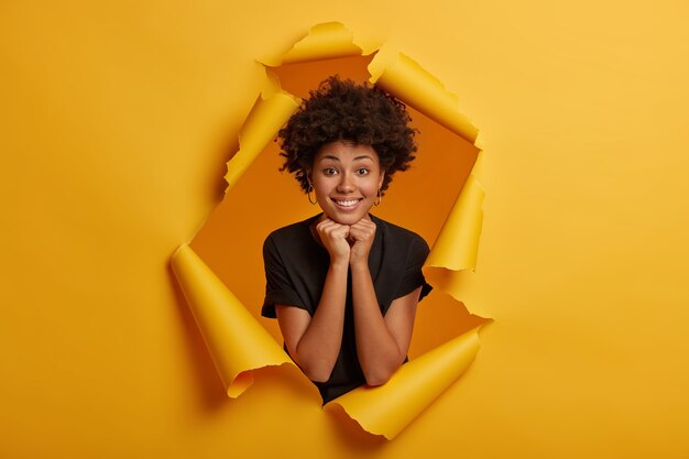 Foto de uma adorável jovem afro-americana sorrindo agradavelmente para a câmera, mantendo as duas mãos sob o queixo, mostrando os dentes brancos, vestida com roupas pretas