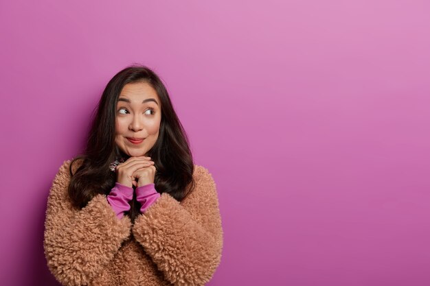 Foto de uma adorável mulher coreana mantendo o olhar de lado, com as mãos embaixo do queixo