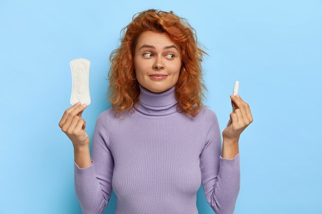 Foto de uma atraente mulher de gengibre segura junta para uso diário e tampão, sugere produtos higiênicos durante dias críticos para as mulheres, escolhe melhor proteção, vestida com blusão casual violeta