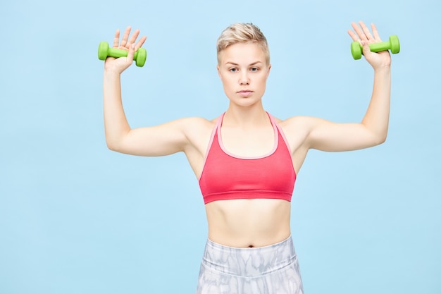 Foto de uma bela jovem esportiva com cabelo de menino, fazendo exercícios físicos, segurando os braços para o lado com dois halteres verdes nas mãos, treinando os músculos do bíceps e dos ombros