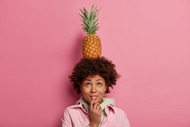 Foto de uma curiosa mulher afro-americana de pele escura carregando abacaxi na cabeça, olhando para cima, vestida com roupas da moda, posa contra uma parede rosa, tem humor positivo, brinca com comida exótica