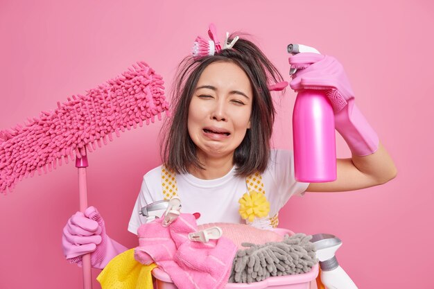 Foto de uma dona de casa estressada chora de desespero, cansada de limpar a casa, segura o esfregão e o detergente tem uma expressão triste. Funciona sobre a casa isolada sobre o fundo rosa. Limpador de fadiga.