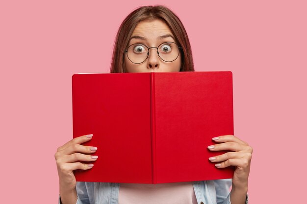 Foto de uma estudante inteligente cobre o rosto com um livro vermelho, olha fixamente com olhos arregalados, fica chocada ao ouvir notícias repentinas, estuda para a prova na faculdade, isolada sobre uma parede rosa