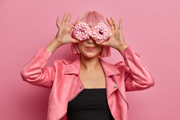 Foto de uma garota asiática elegante de cabelo rosa cobre os olhos com rosquinhas deliciosas, gosta de confeitaria saborosa e aromática, come rosquinhas com cobertura