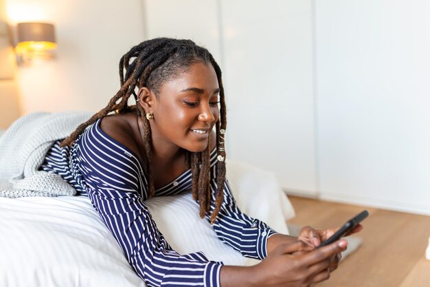 Foto de uma jovem africana sorridente tem uma videochamada à noite enquanto está deitada em uma cama em seu quarto