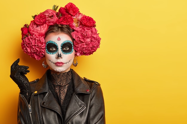 Foto de uma jovem com o rosto pintado de maneira brilhante para lembrar crânios, usa uma jaqueta de couro preta e luvas e usa uma guirlanda feita de flores vermelhas aromáticas