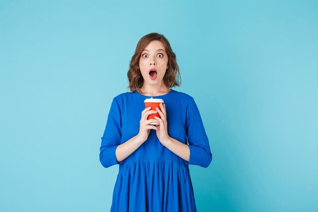 Foto de uma jovem de vestido azul em pé com uma xícara de café para ir e olhando espantada para a câmera sobre fundo azul