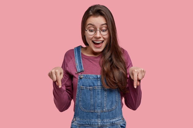 Foto de uma jovem europeia atraente e alegre, com cabelos lisos e escuros, aponta para baixo, tem uma expressão feliz, vestida com roupas casuais