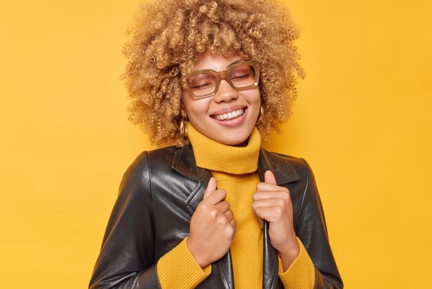 Foto de uma jovem europeia positiva com cabelos cacheados, vestida com roupas elegantes, lembra boas lembranças, mantém os olhos fechados, isolados sobre fundo amarelo. Conceito de roupas e emoções de pessoas