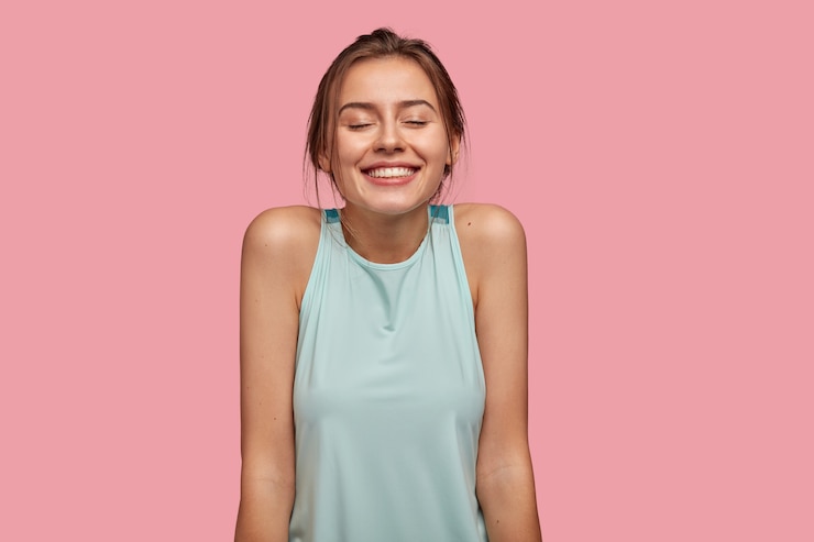 Foto grátis foto de uma jovem europeia satisfeita com um sorriso largo e agradável, mantém os olhos fechados, sonha com algo agradável, mostra os ombros nus, vestida com colete casual, isolada sobre uma parede rosa