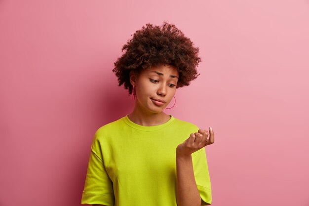 Foto de uma jovem impressionada olha para sua nova manicure, não gosta de unhas polidas, vestida com uma camiseta verde brilhante, isolada na parede rosa. Senhora olha atentamente para os dedos