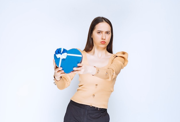 Foto de uma jovem modelo segurando uma caixa de presente em forma de coração contra uma parede branca.
