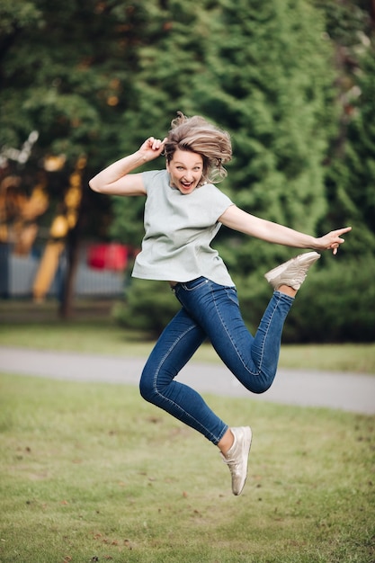 Foto de uma jovem mulher branca com cabelo louro em uma camiseta azul, jeans e tênis salta e se alegra