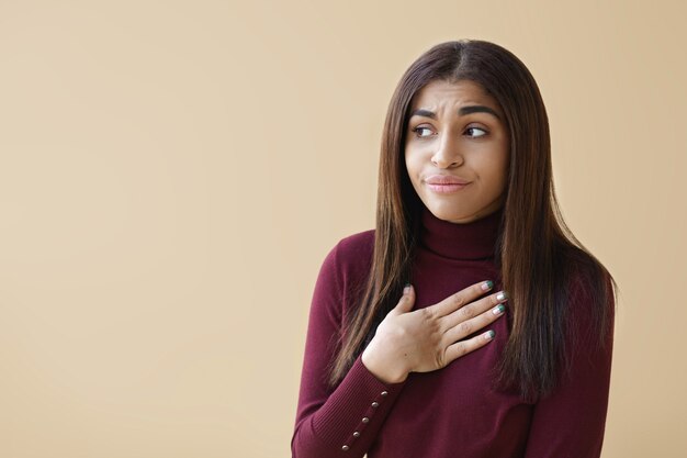 Foto de uma jovem mulher de pele escura chateada, usando um pescoço de tartaruga roxo, desviando o olhar com uma expressão de desgosto, mantendo a mão no peito como se tivesse um mau pressentimento sobre a tomada de alguma decisão importante