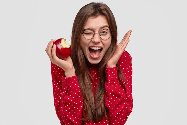 Foto de uma jovem satisfeita pisca os olhos, levanta a mão perto da cabeça, morde maçã fresca e tem uma expressão alegre, vestida com uma blusa de bolinhas vermelhas