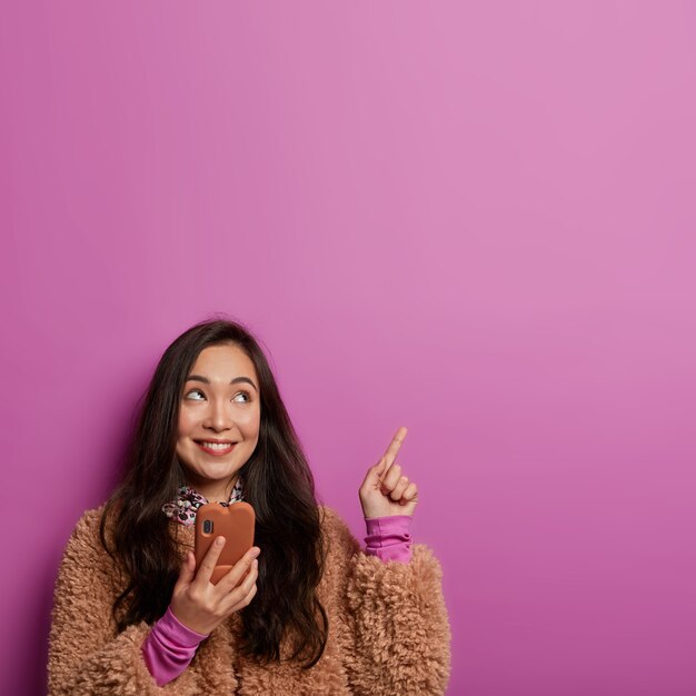 Foto de uma jovem sonhadora com sorriso dentuço, cabelo escuro, tem uma ideia interessante, recomenda algo no espaço em branco, usa o celular