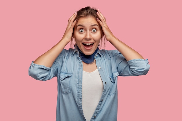 Foto de uma linda garota espantada com expressão facial surpresa e feliz, mantendo as duas mãos na cabeça, não posso acreditar no sucesso, vestida com uma camisa jeans da moda, isolada na parede rosa