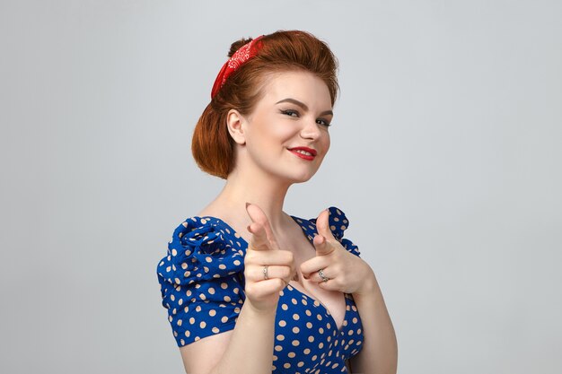 Foto de uma linda jovem europeia ruiva com maquiagem brilhante e penteado estiloso, olhando para a câmera com um sorriso amigável, apontando os dois dedos indicadores, fazendo a escolha certa, escolhendo você