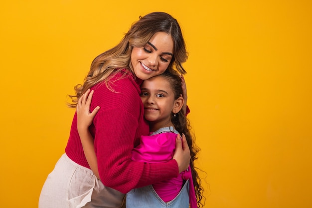 Foto de uma linda jovem mamãe segurando seus braços abraçando sua filhinha bom humor adorável coração sincero sentimentos nas costas camisetas jeans isoladas com fundo de cor amarela