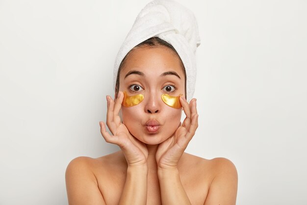 Foto de uma linda modelo feminina aplica almofadas amarelas sob os olhos para reduzir as rugas, tem procedimentos anti-envelhecimento, mantém os lábios dobrados, fica sem camisa dentro de casa, toalha enrolada na cabeça. Conceito de cosmetologia