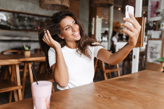 Foto de uma linda mulher bronzeada tirando uma selfie em um café