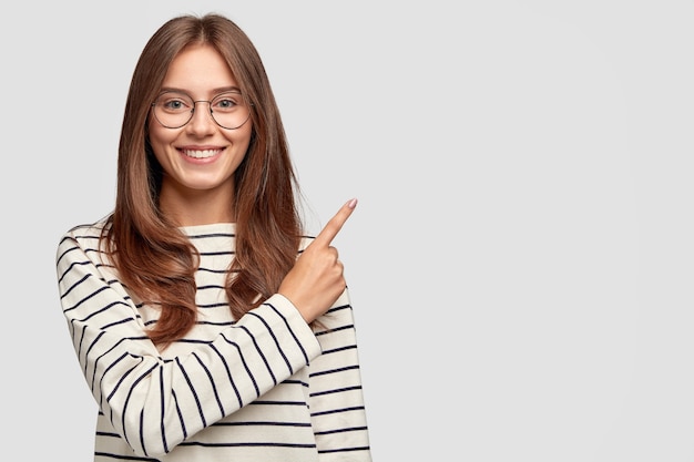 Foto de uma linda mulher caucasiana com expressão alegre, aponta com o dedo indicador para o espaço em branco da cópia, vestida com suéter listrado, mostra espaço livre no canto superior direito para sua promoção