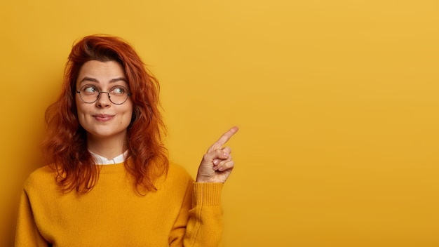 Foto de uma linda mulher ruiva aponta o dedo indicador para o lado, demonstra promo à direita, parece com uma expressão interessante, tem cabelo ruivo ondulado