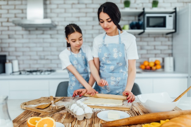 Foto de uma massa e ingredientes na mesa da cozinha e duas meninas cozinhando