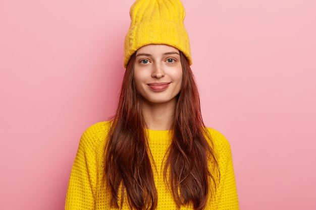 Foto de uma modelo feminina bonita tem longos cabelos escuros, olha diretamente para a câmera, usa um chapéu amarelo vívido e uma camisola de malha, estando de bom humor, isolada sobre um fundo rosa.