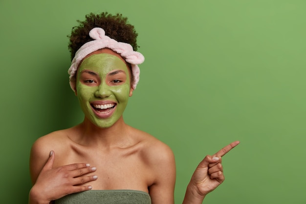 foto de uma mulher afro-americana de aparência agradável e alegre aplica máscara de beleza no rosto, recomenda produto de cosmetologia, indica no espaço da cópia, isolado na parede verde. Conceito de cuidados com a pele