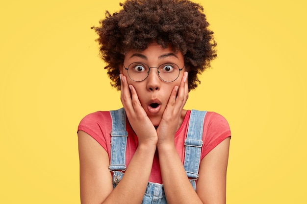 Foto de uma mulher afro-americana surpresa que toca as bochechas, abre os olhos e a boca amplamente, vestida com roupas casuais, isolada sobre a parede amarela. Mulher chocada de raça mista posa sozinha dentro de casa.