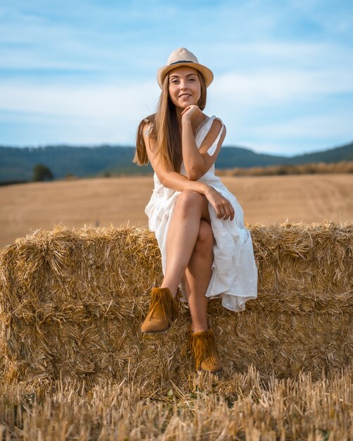 Foto de uma mulher atraente em um vestido branco sentada em um galho de feno e olhando para a câmera