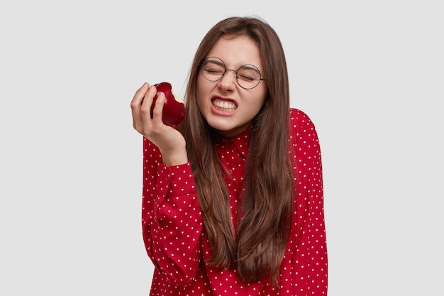 Foto de uma mulher bonita aperta os dentes, morde maçã vermelha fresca, vestida com uma camisa elegante, usa óculos