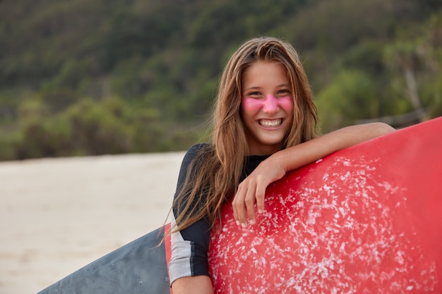 Foto de uma mulher bonita com surf zinco para proteção facial, segurando uma prancha de surf encerada, poses sobre um penhasco