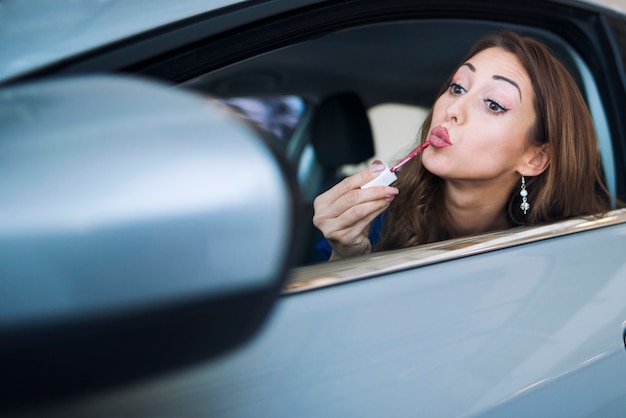 Foto de uma mulher bonita sentada no carro, olhando para o espelho retrovisor, passando batom e aplicando maquiagem