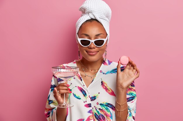 Foto de uma mulher de pele escura satisfeita fecha os olhos e sorri suavemente gosta de passar o tempo em casa segurando um delicioso macaroon e copos de coquetel de martini usa roupão isolado sobre a parede rosa