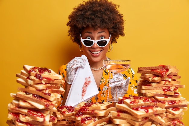 Foto de uma mulher de pele escura sendo arrumada e bem vestida, posa com copo de coquetel e guardanapo, usa roupas estilosas, passa o tempo livre em restaurante de luxo, deliciosas torradas de geleia em primeiro plano