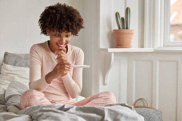 Foto de uma mulher étnica sorridente e feliz olha felizmente para o resultado positivo do teste de gravidez, não consigo acreditar que ela se tornará mãe, senta-se na cama, sente-se satisfeita, aproveita o tempo de recreação, antecipa o bebê