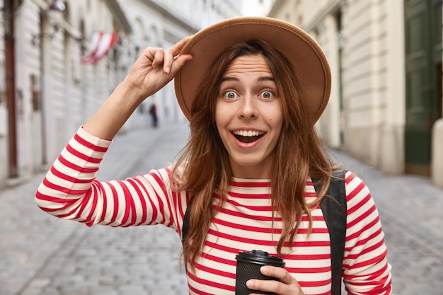 Foto de uma mulher europeia alegre e surpresa segurando o chapéu, bebe café para viagem, caminha na rua da cidade