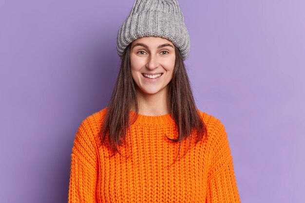 Foto de uma mulher europeia atraente com cabelo escuro comprido sorriso agradável feliz por ter as férias de inverno usa chapéu e suéter de malha laranja.